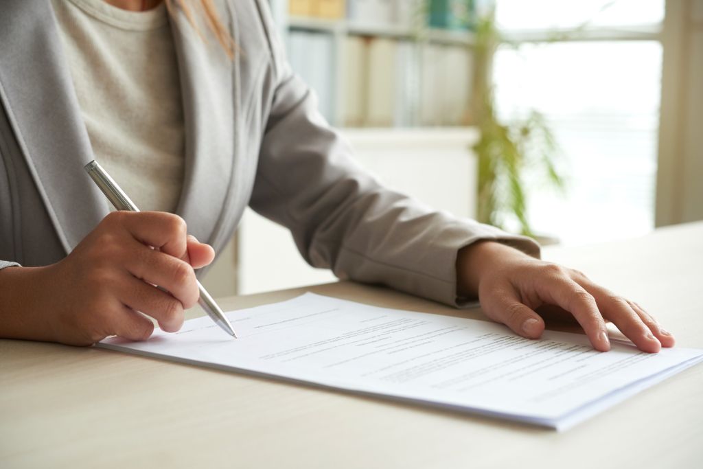 Dłonie kobiety trzymające długopis w prawej ręce, podpisuje dokument, który lezy na biuru przed nią.